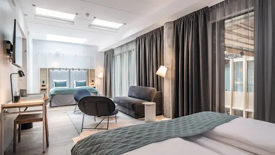 Ett modernt rum med en dubbelsäng och teal-sängkläder, en soffa och ett skrivbord, omgiven av stora fönster och gardiner, upplyst av taklampor.