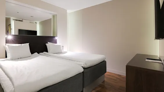 Två prydligt bäddade enkelsängar står med en sänggavel, upplysta av läslampor, i ett modernt, minimalistiskt hotellrum med trägolv och en väggmonterad TV.