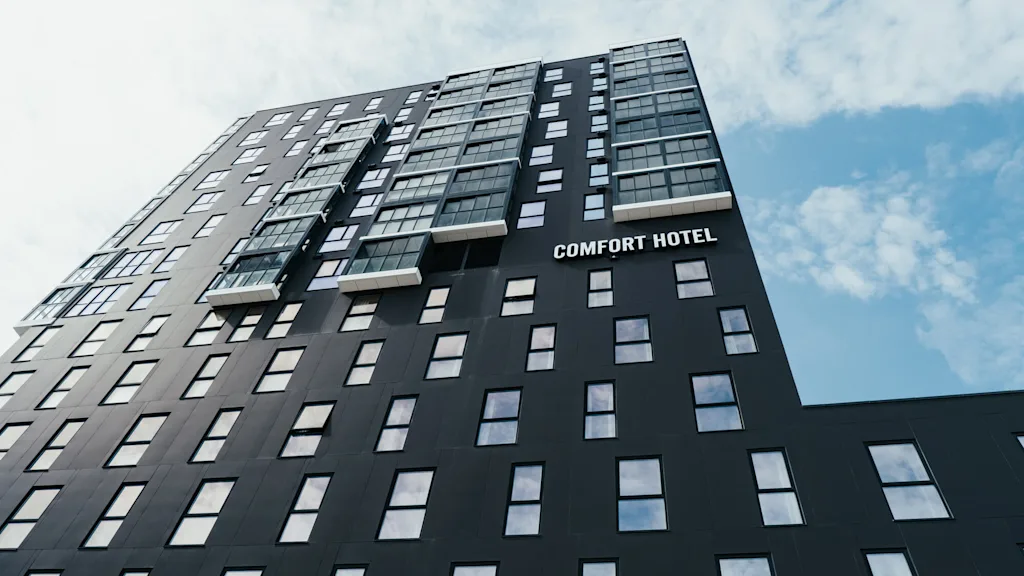 En høj, moderne hotelbygning med mange vinduer, mærket "COMFORT HOTEL," står mod en lys, skyet himmel.