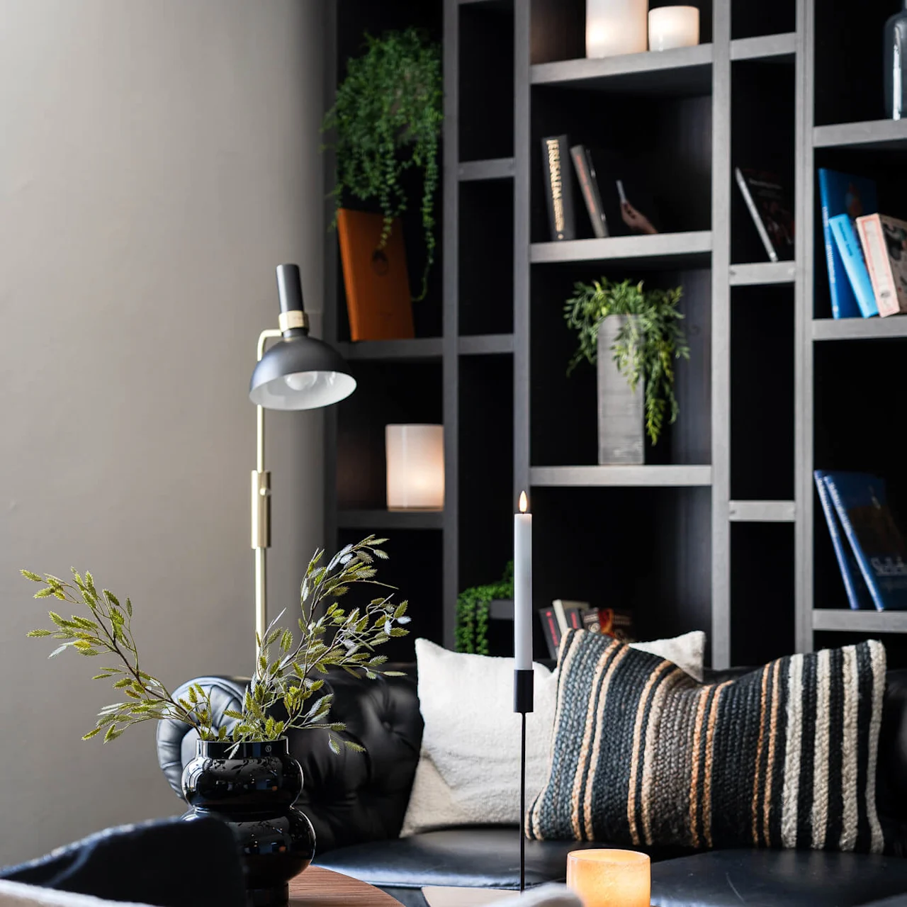 En svart lädersoffa med kuddar och en växt står framför mörka hyllor fyllda med böcker och ljus. En modern lampa och tänt ljus ger stämning.