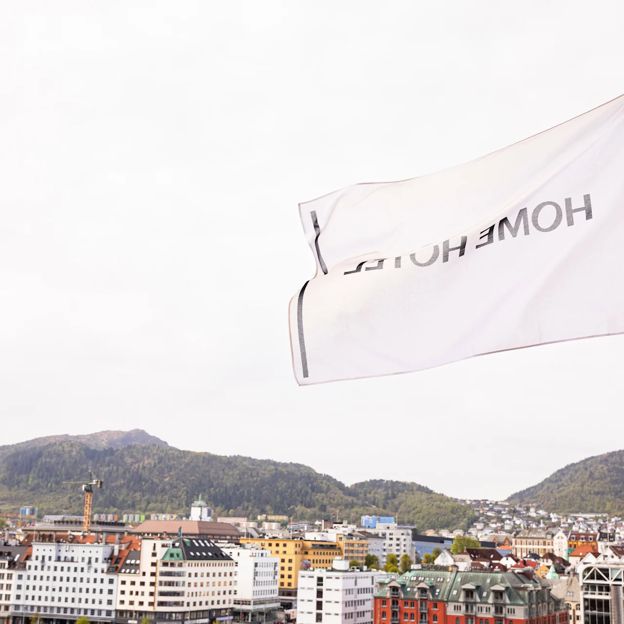 A white flag reads "HOME HOTEL" while fluttering in the wind above a cityscape backdrop with colorful buildings and distant green hills.