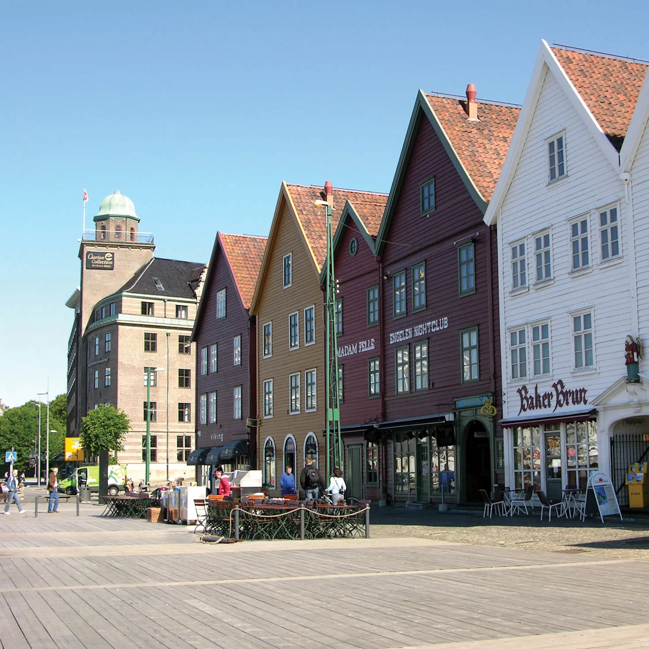 Historic colorful wooden buildings line a cobblestone square with outdoor seating and pedestrians, under a clear blue sky.