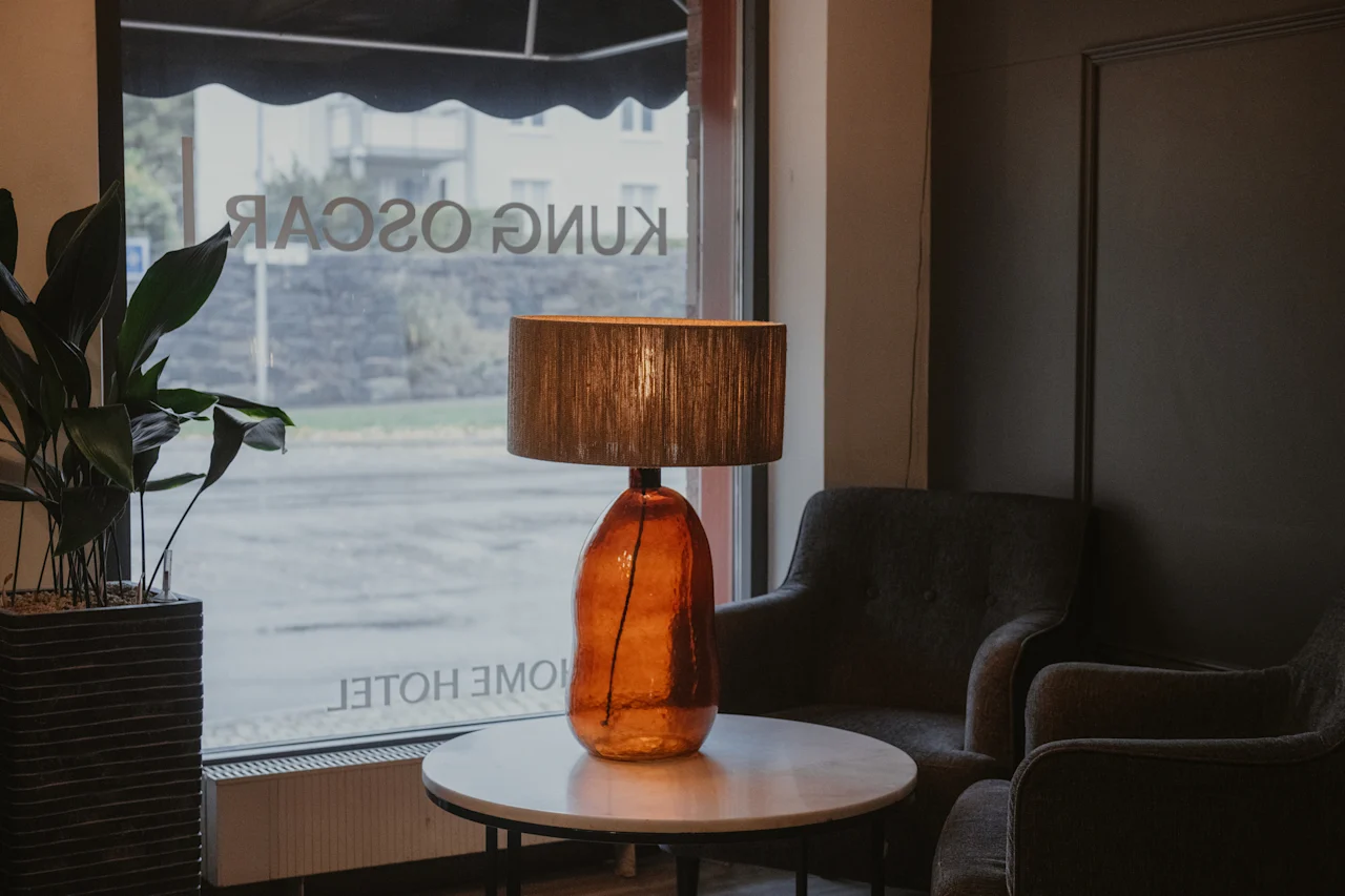 A welcoming hotel lounge with a warm amber lamp, comfortable armchairs, and a plant by a window at Home Hotel Kung Oscar.