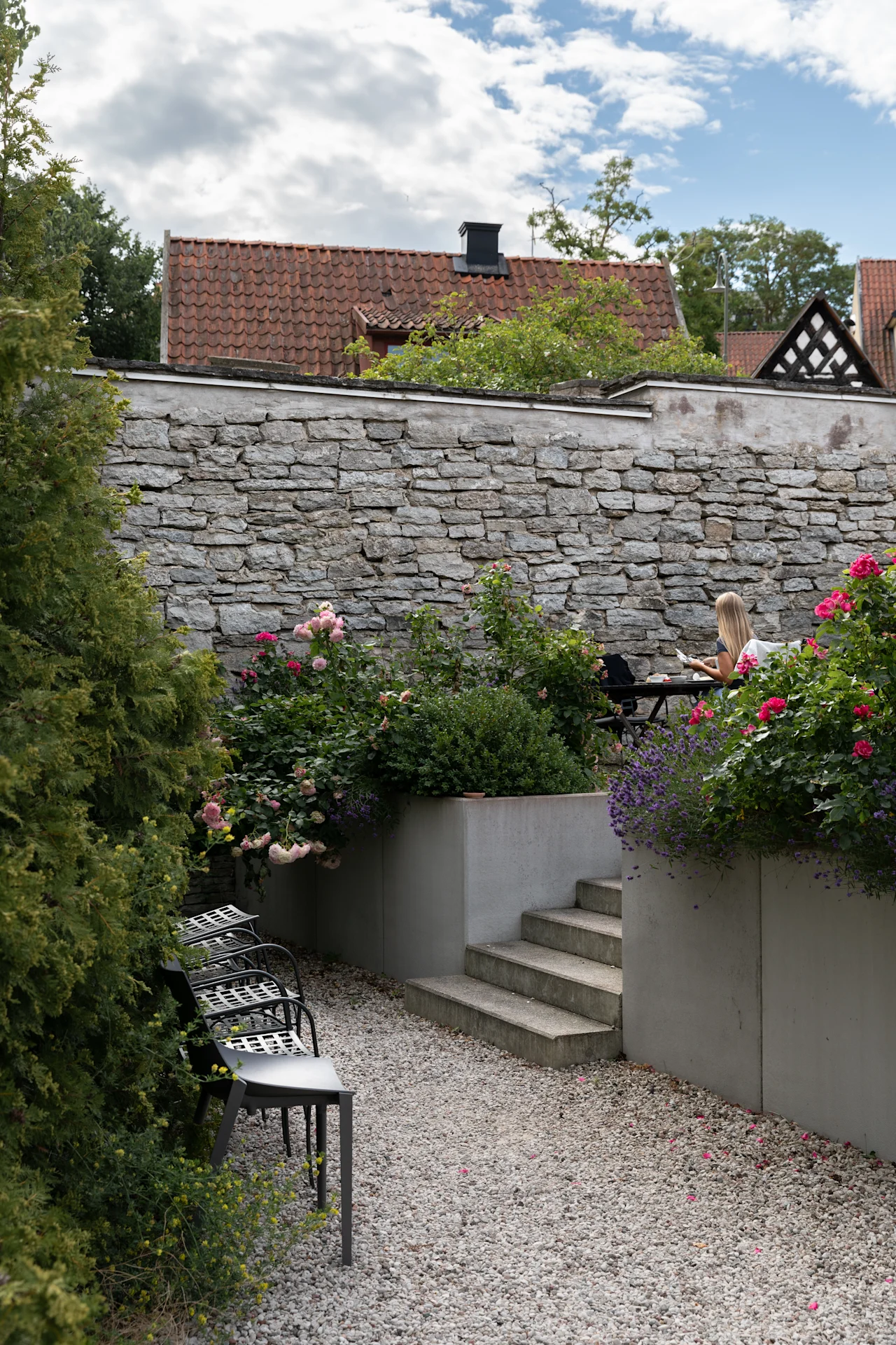 En kvinde sidder ved et bord og læser i en have med blomstrende blomster. Stentrapper og murer omgiver hende, og tagene er synlige under en delvist overskyet himmel.