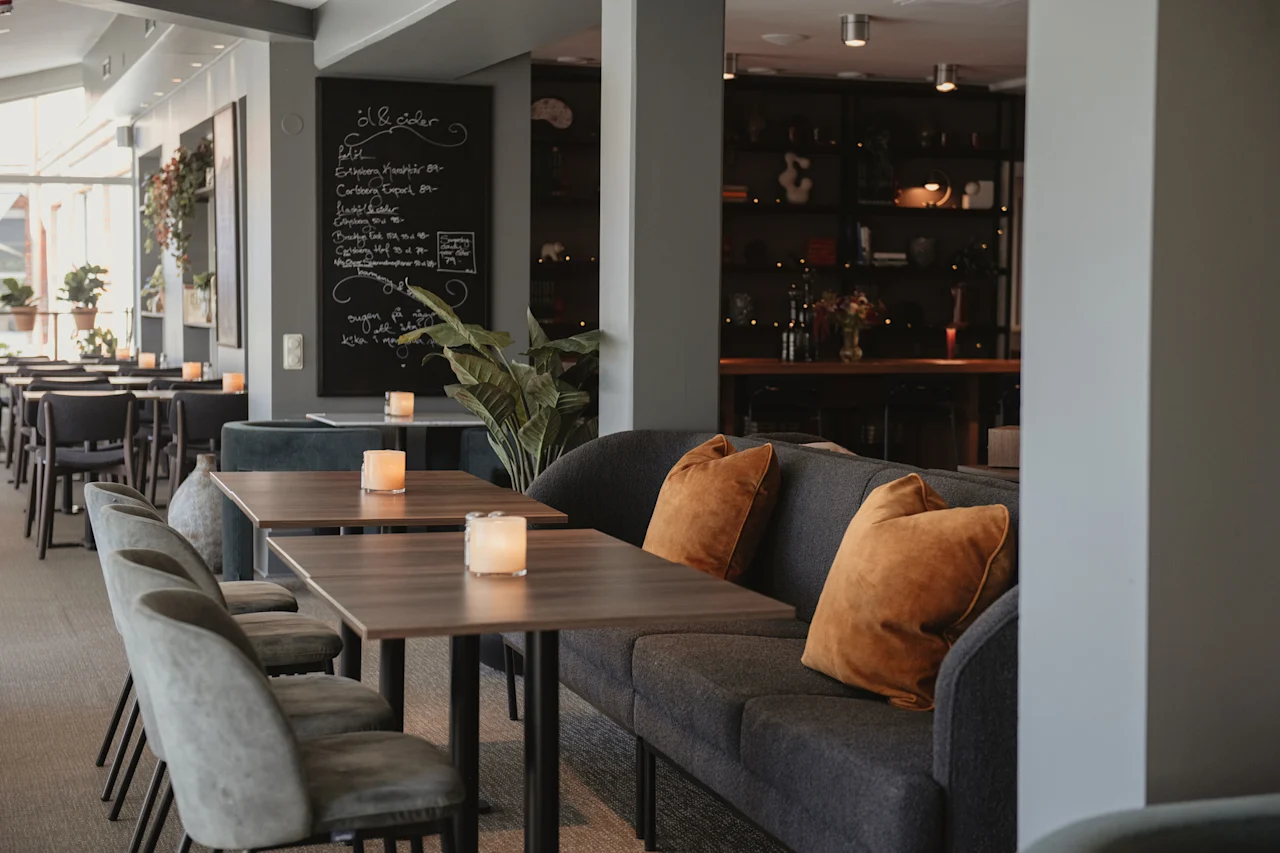 A cozy restaurant with tables, comfortable seating, and a menu board displaying drinks like Ol & cider, Pilsner, Cofee, Te, Vin.