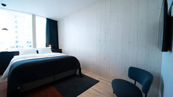 Bed sits against a white wooden wall, covered with black and white bedding. Next to it, a chair with a black padded seat and backrest. The room is well-lit by a large window and ceiling lights.