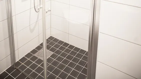En duschmunstycke är monterat på en vägg i ett kaklat hörn av badrummet, med vita väggar och ett mörkt mönstrat golv, omgivet av glasdörrar.