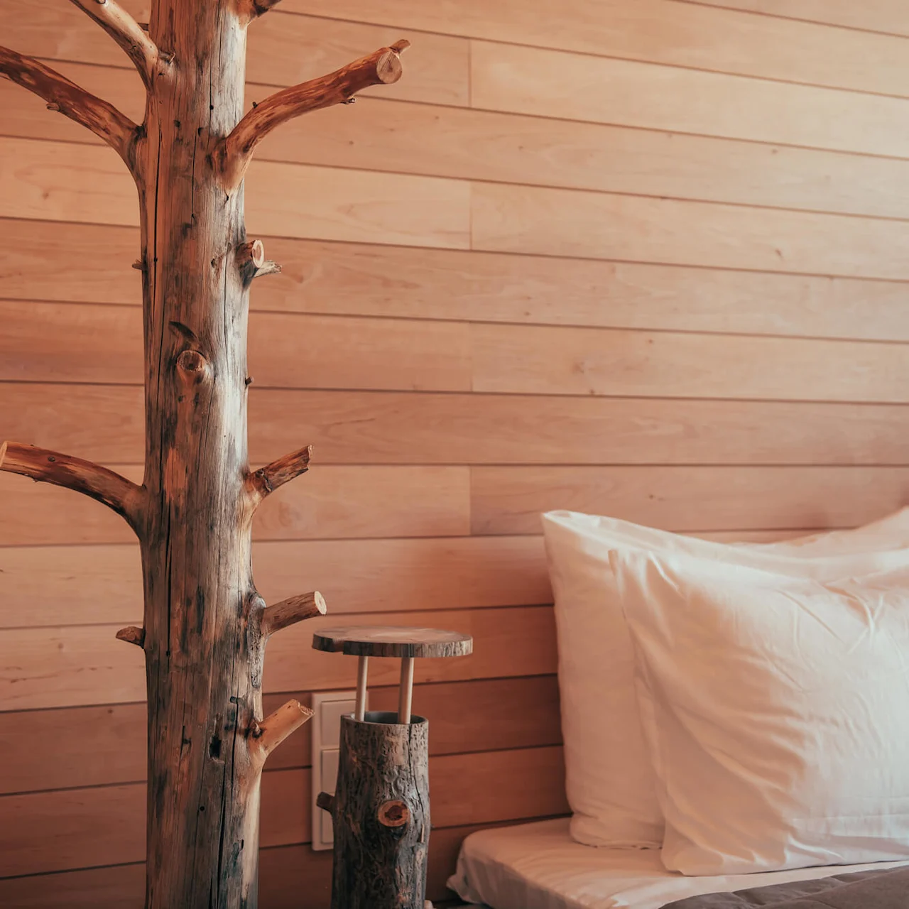 Wooden tree trunk serves as decorative element beside a bed with white pillows, set against a wooden paneled wall in a cozy room.