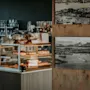 Et glasmontre indeholder et udvalg af bagværk under glaskupler. I nærheden pryder sort-hvide fotografier en trævæg. Miljøet ser ud til at være en hyggelig café.