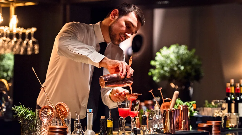 Baarimikko kaataa kirkkaanpunaista cocktailia siivilän läpi lasiin. Ympäröity pulloilla ja baarityökaluilla himmeästi valaistulla baaritiskillä.