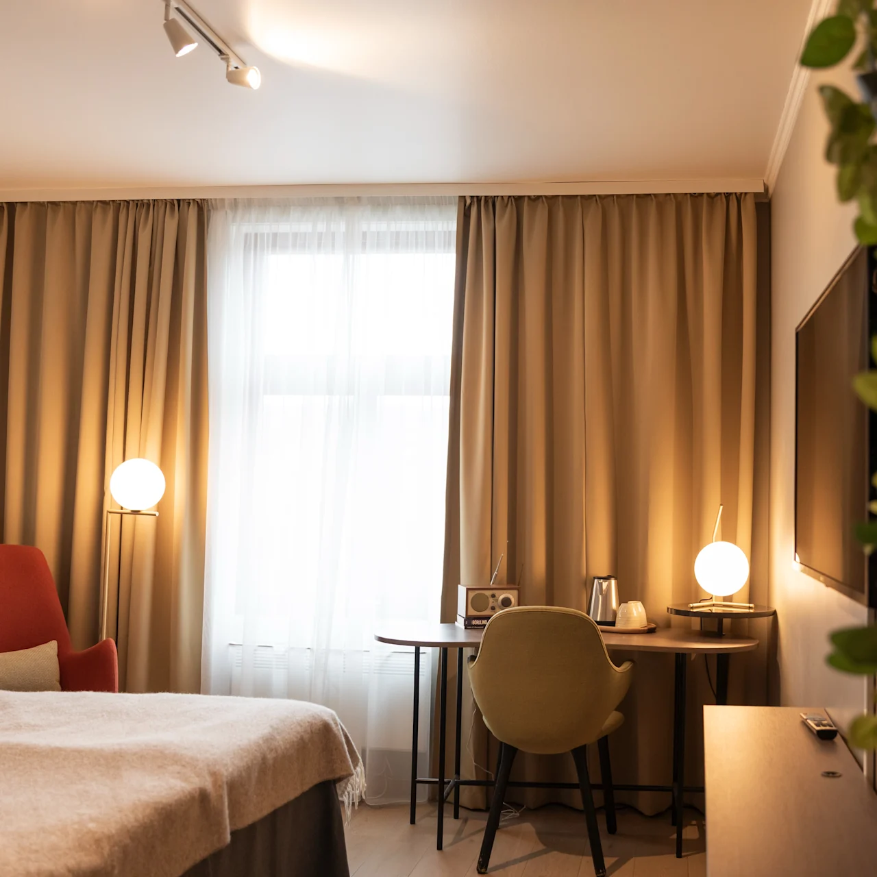 Ett prydligt arrangerat hotellrum har en säng och en skrivbordsstol. Varm belysning lyser upp ett delvis förhängt fönster, med en TV och en bladväxt på väggen.