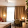 Et pent organisert hotellrom har en seng og en skrivebordstol. Varm belysning lyser opp et delvis dekket vindu, med en TV og en grønn plante på veggen.