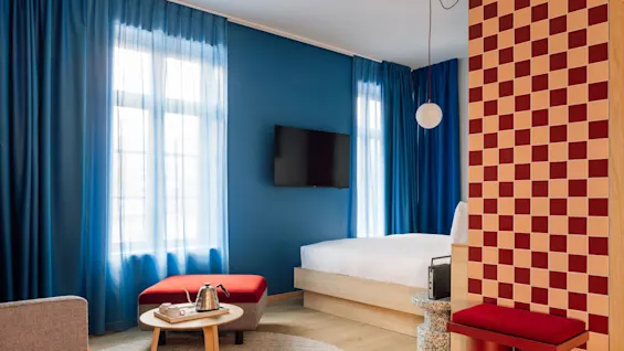 A modern hotel room with blue curtains, a bed, a TV, and a seating area. Red and beige checkered patterns on walls and floor.