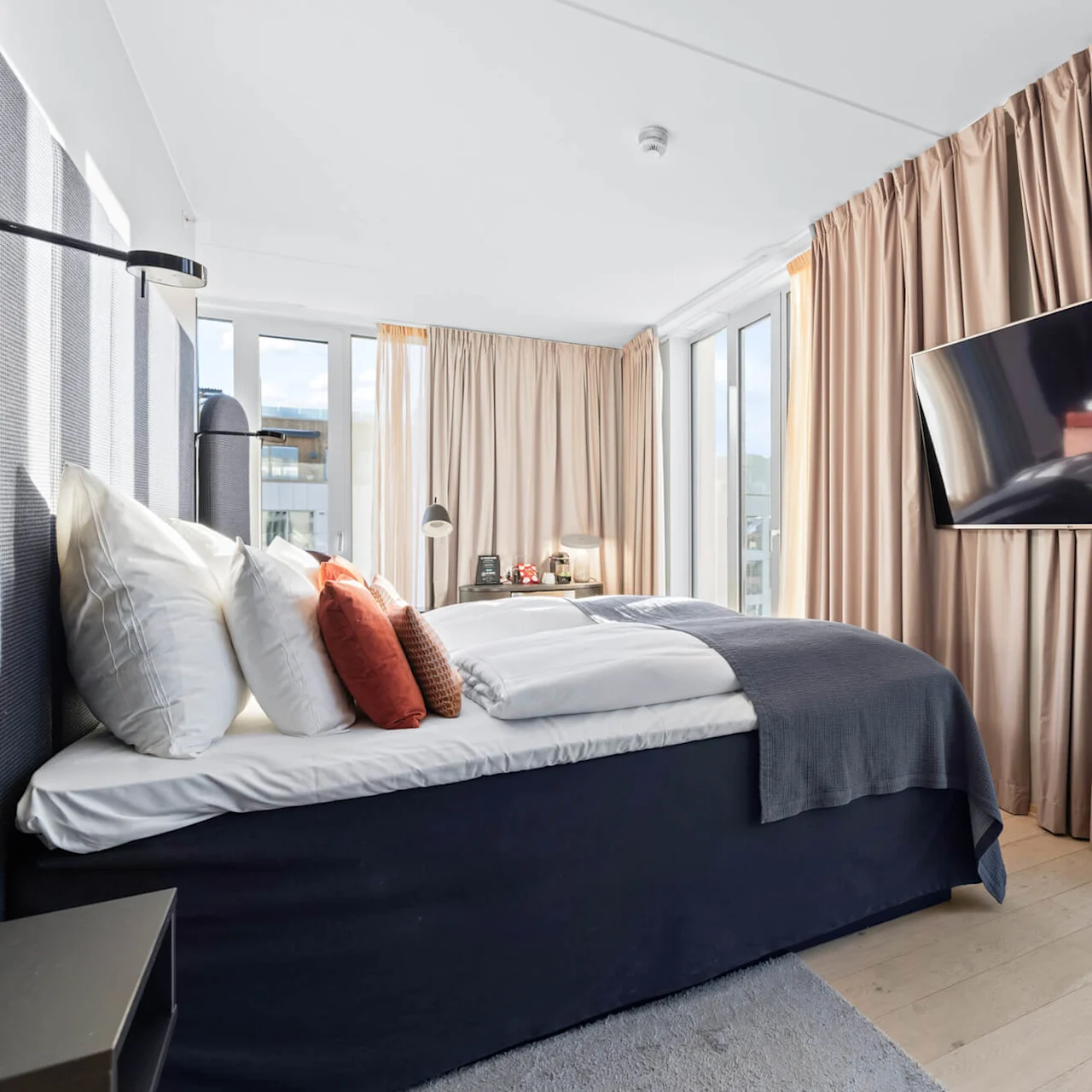 A bed with multiple pillows is positioned near large windows, with beige curtains and a wall-mounted TV. Two bedside lamps are on a table, illuminating the modern bedroom.