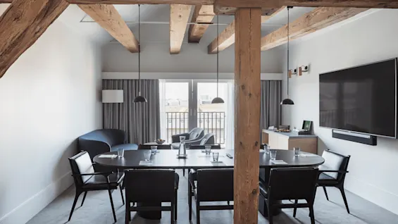 Meeting room with a large oval table surrounded by chairs, set with glasses and papers. Rustic wooden beams overhead and a large TV screen on the wall.