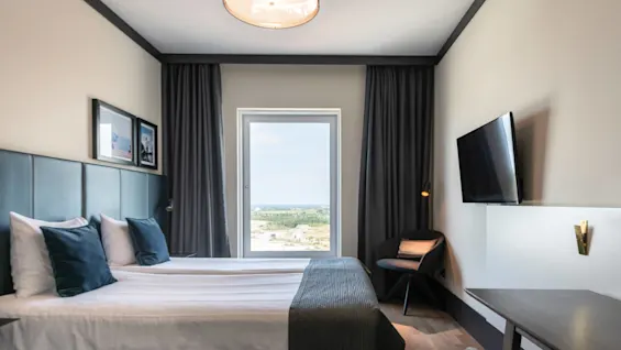 A neatly made bed with pillows rests beside a large window, revealing a scenic landscape. A wall-mounted TV faces a nearby chair and desk in a modern room.