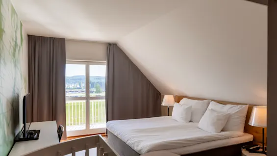 Et lyst hotelværelse med en seng, et skrivebord, et TV og en balkon med udsigt over et grønt landskab. En babyseng er i forgrunden.