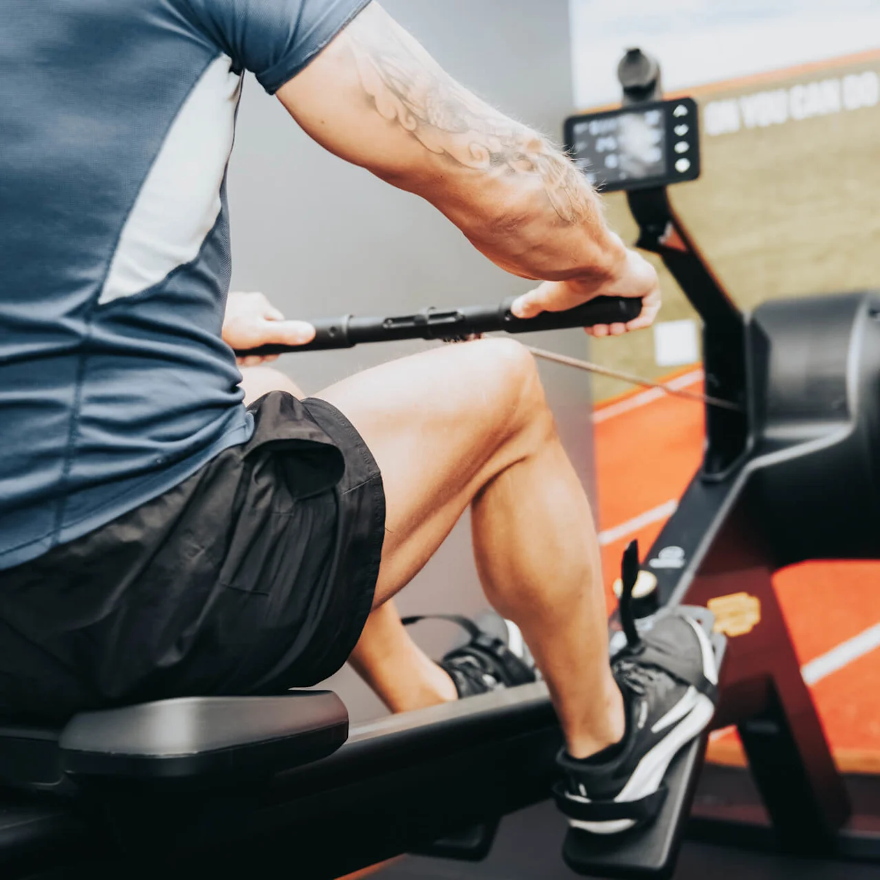 A person sits on a rowing machine, pulling the handle, wearing athletic gear. A motivational sign in the background reads, “YOU CAN DO IT!"