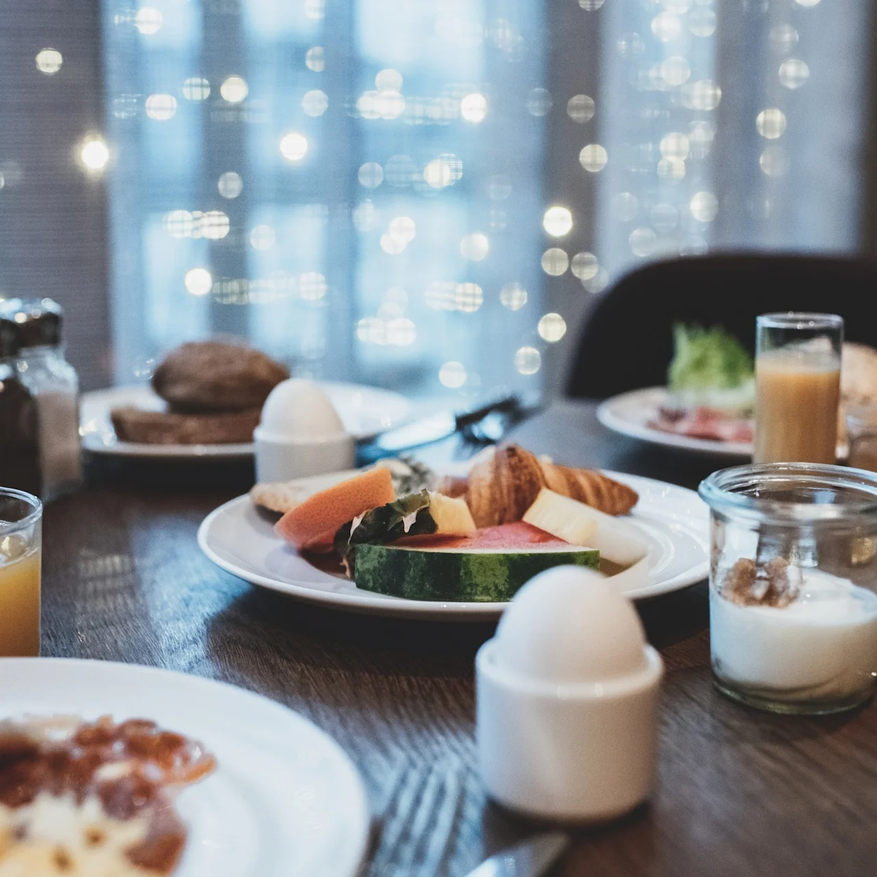 Ett frukostbord dukat med tallrikar med frukt, ägg och croissanter, omgivna av drycker och kryddor, i ett varmt upplyst rum med suddiga ljus utanför ett fönster.