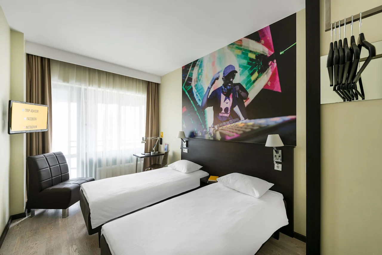 Two neatly made single beds face a wall mural of a DJ in a modern hotel room. A television displays options: "TRIP ADVISOR," "FACEBOOK," and "INSTAGRAM." A chair and desk with lamp are nearby.