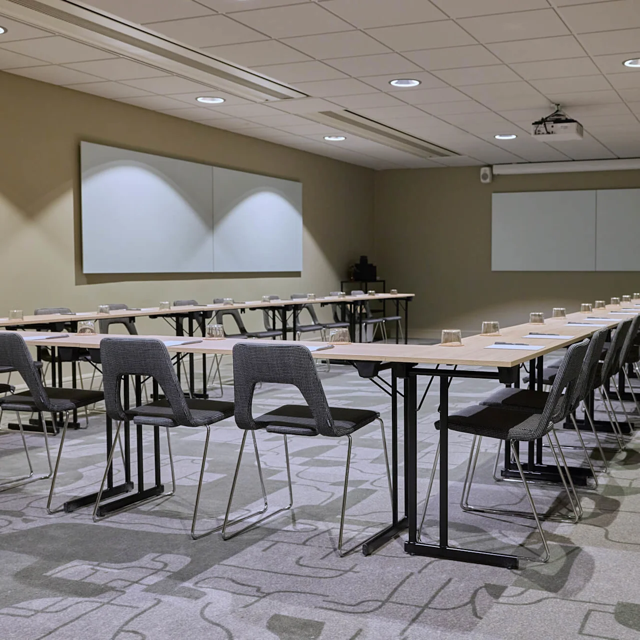 Meeting room with rectangular tables arranged in a U-shape, chairs, notepads, and glasses set. Neutral-toned walls and ceiling with spotlights surround the space, featuring two whiteboards and a potted plant.