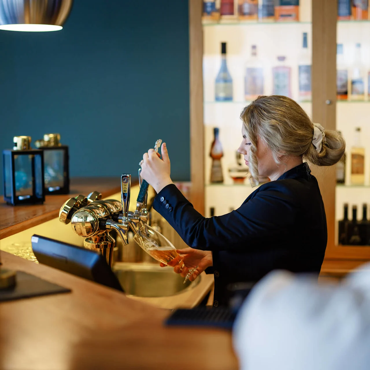 Bartender häller upp öl från en kran i ett glas, på en bar med en trädisk och flaskor i bakgrunden.