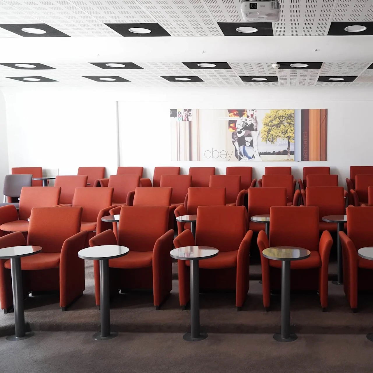 Rows of red chairs with individual round tables in a lecture hall; overhead projector and abstract paintings on the walls; ceiling with patterned panels features circular lighting. Text: "OBEY" and "SCAPE."  