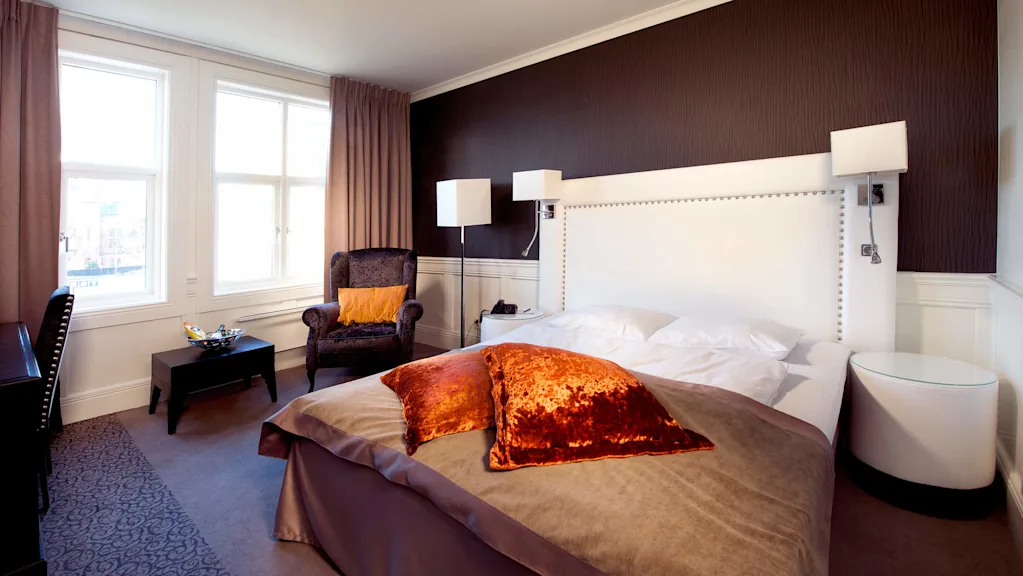 A neatly made bed in a warmly lit elegant bedroom, with an upholstered chair and a window with curtains.