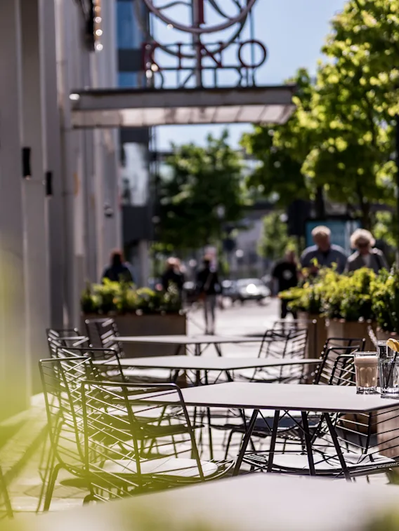 Utendørs metallkafébord med glassdrikker, omgitt av grønne planter og noen få mennesker som går langs en solbelyst gate under en delvis skyet himmel.