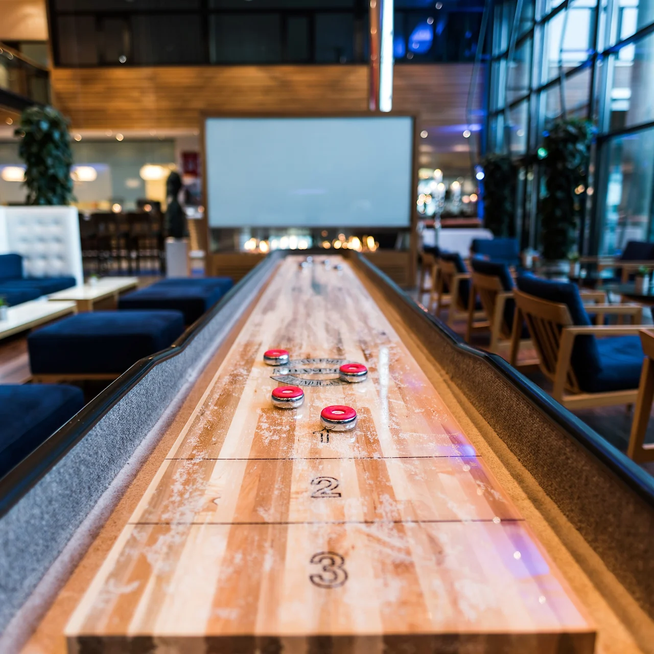 Et shuffleboardbord med røde brikker står i en moderne lounge med blå polstring, bord og store vinduer.