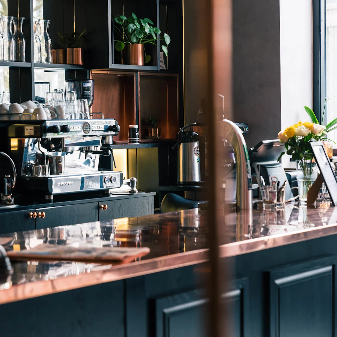 En kaffebar viser en moderne espressomaskin, omgitt av kopper og glass. I nærheten dekorerer planter og blomster disken, og skaper en innbydende atmosfære.