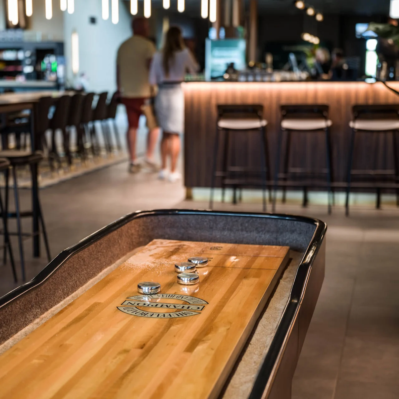 Shuffleboard med metallpucker hviler på en treoverflate, omringet av en moderne barinnstilling med høye stoler og folk som snakker i bakgrunnen.