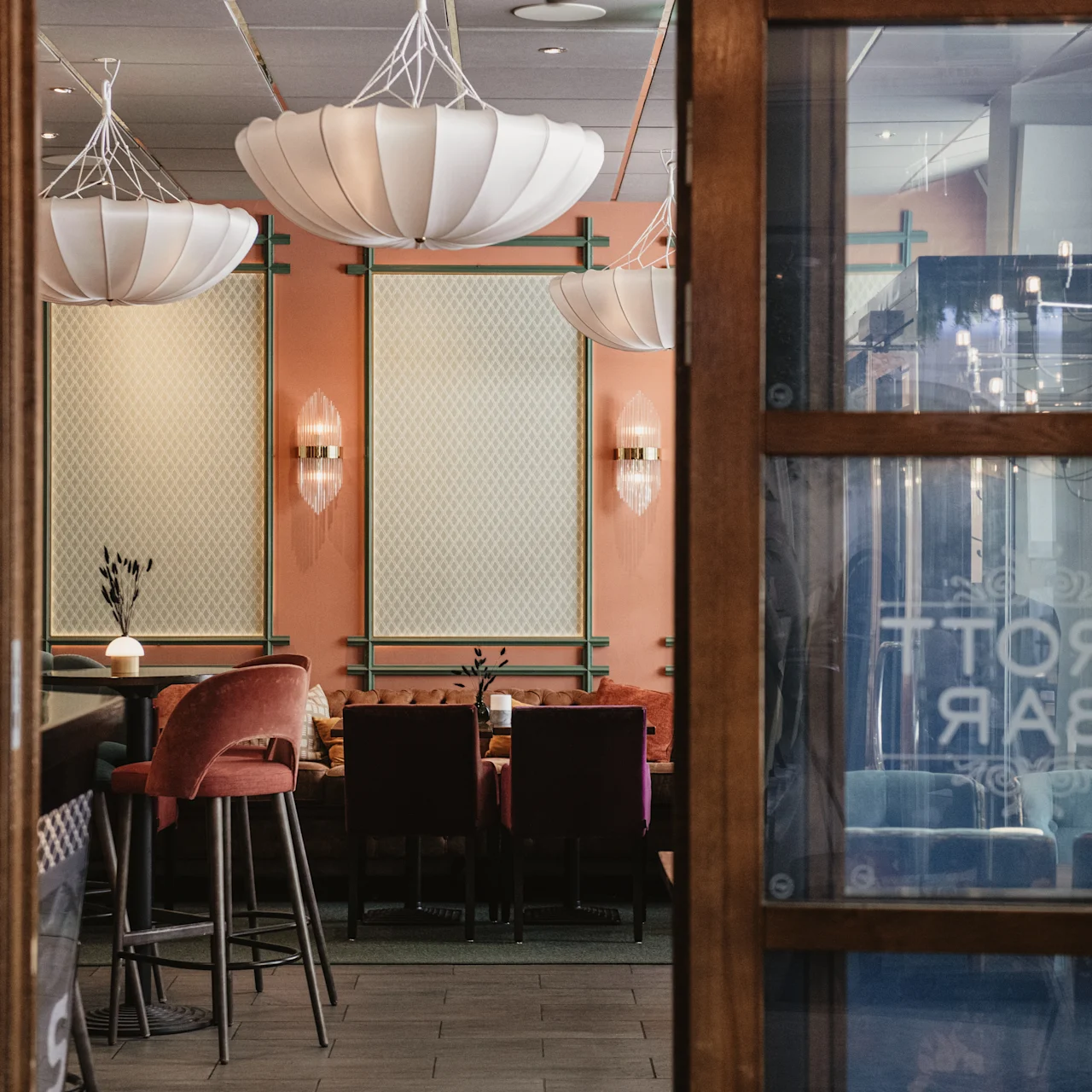 Et spiseområde med elegante hvide pendellamper hænger over borde omgivet af stole. Lyserøde vægge med indrammet kunst skaber en hyggelig atmosfære. Døren siger "DROTT BAR".