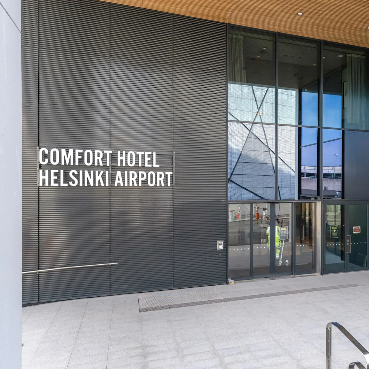 Comfort Hotel Helsinki Airport indgang har glasdøre og høje vinduer. Tekst på bygningen lyder "COMFORT HOTEL HELSINKI AIRPORT". Omgivelser: moderne arkitektonisk design.