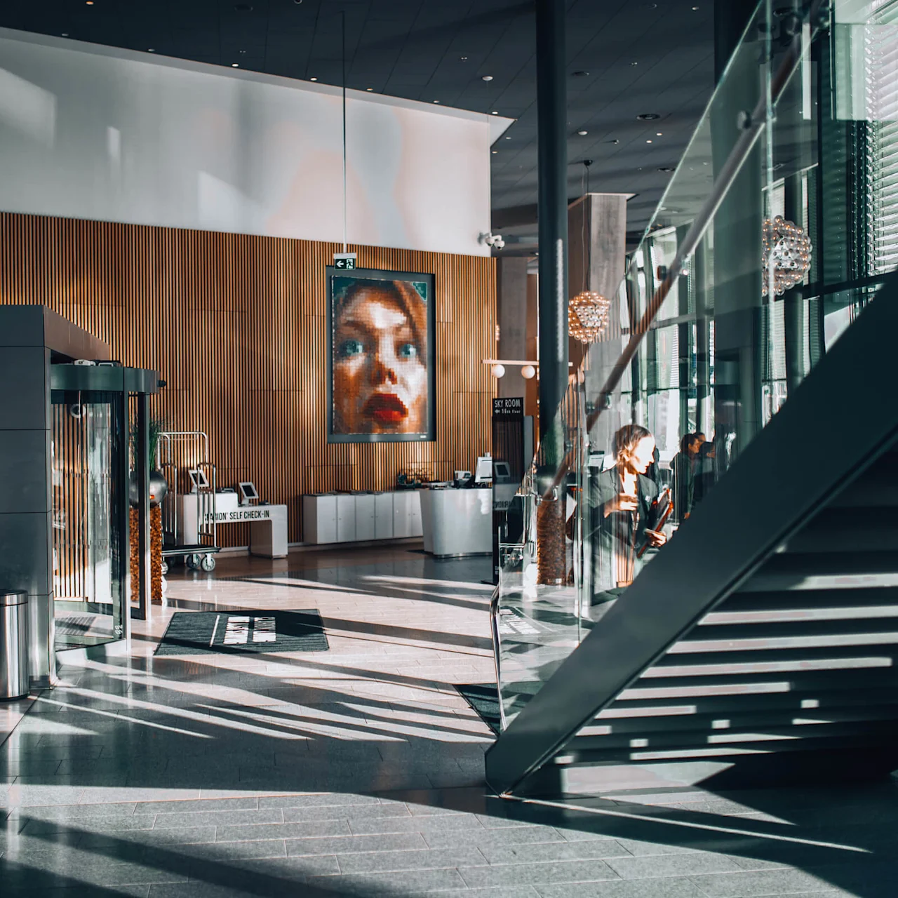 Et rummeligt, moderne lobbyområde har et stort portræt på en trævæg; personer slapper af ved glastrapper. Teksten lyder, "SKY ROOM SELF CHECK-IN" og "SKY ROOM".