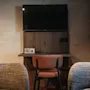 A hotel room corner with a wall-mounted TV above a desk, a chair, and two armchairs, ready for use.