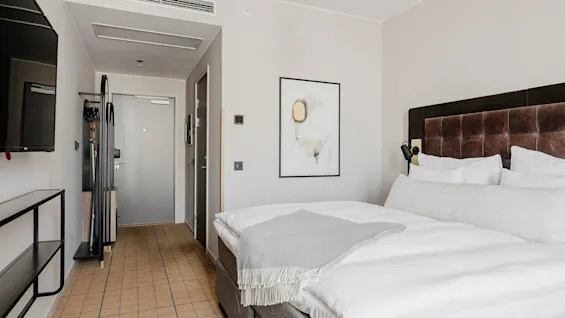 En pænt redt hotelseng med hvidt sengetøj og et gråt tæppe er placeret mod en brun polstret sengegavl. Et moderne, minimalistisk rum med en vægmonteret TV og abstrakt kunst.