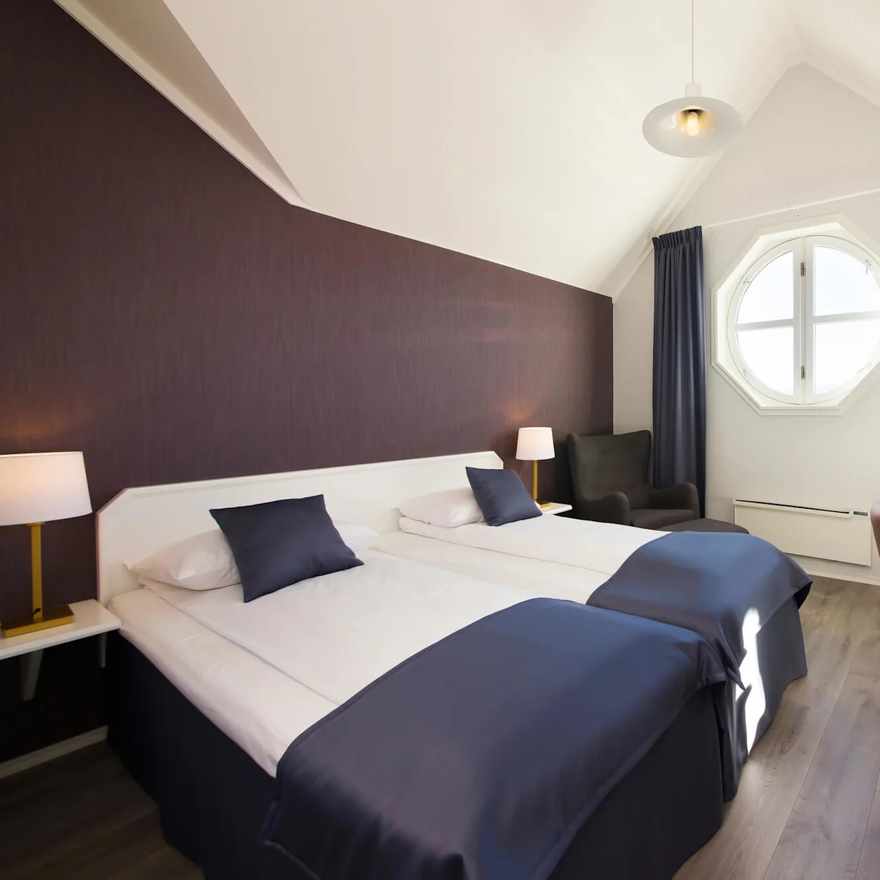 Ett välbelyst hotellrum med två enkelsängar, stolar, bord och ett åttakantigt fönster, som visar en ren, modern design.