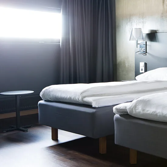 To pent redde enkeltsenger med hvitt sengetøy står side om side i et moderne hotellrom med mørke gardiner og tregulv, under myk belysning.