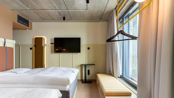 Et moderne hotelværelse med to pænt redte senge, et vægmonteret TV og en bænk ved vinduet. Miljøet er minimalistisk og lyst.