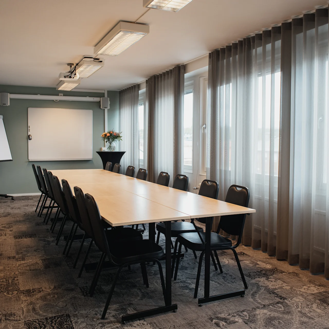 A long conference table surrounded by black chairs sits in a well-lit meeting room. Windows with sheer curtains line one side, and a flip chart and projector screen are visible.