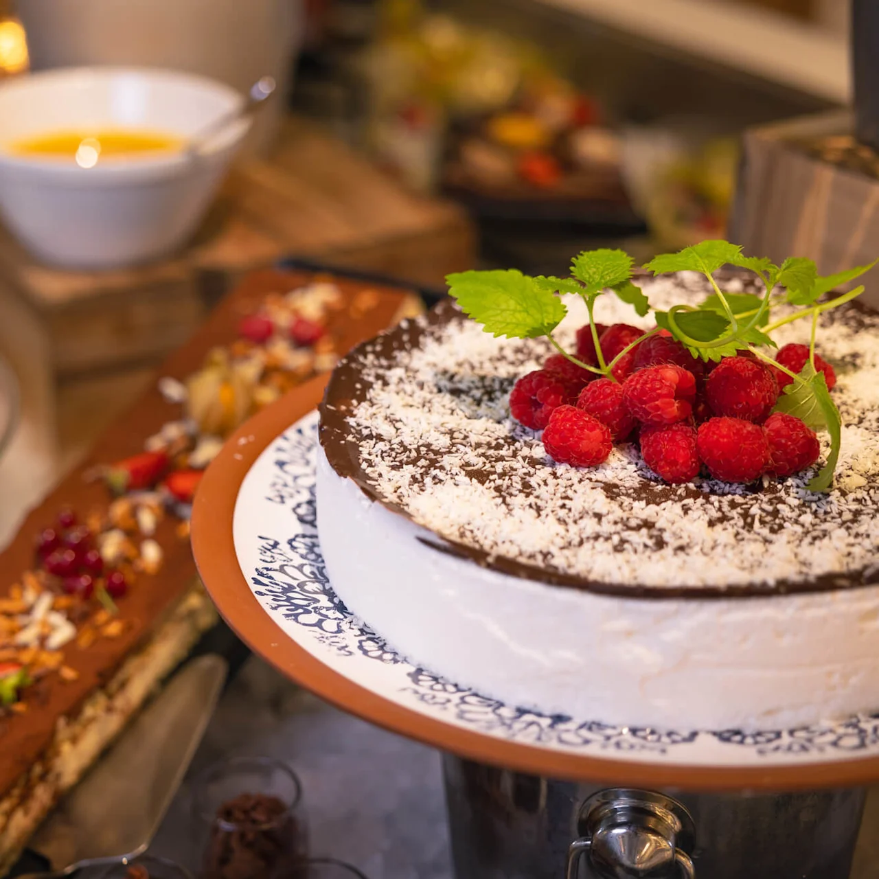 En kake toppet med bringebær og mynte, drysset med kokos, står på et fat. I nærheten er andre desserter, inkludert tiramisu, på et buffébord.