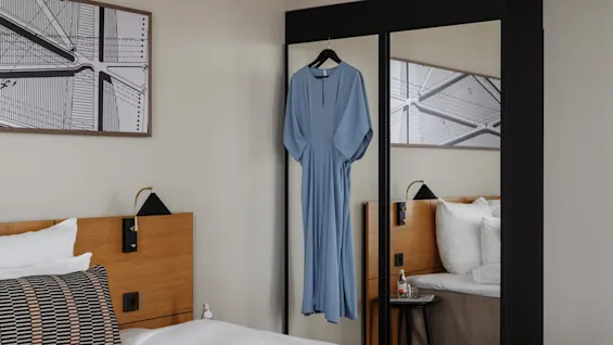 Et hotellrom med en seng og et garderobeskap. En blå kjole henger på den speilvendte garderobedøren, klar for dagen.