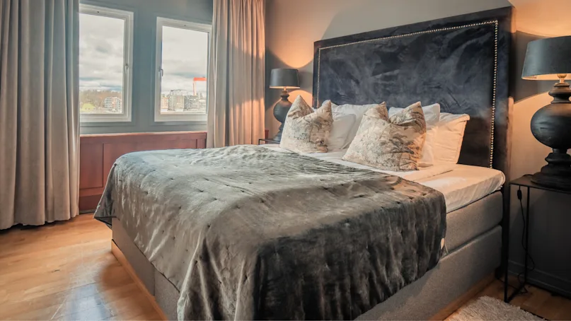 A bed with gray bedding is neatly arranged, topped with decorative pillows, situated in a warmly lit bedroom with wooden floors and a large window showing an urban view.