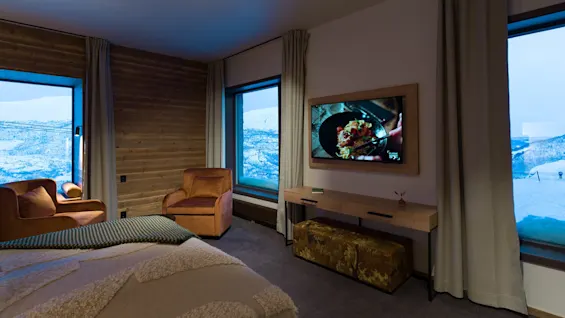 A comfortable room features two brown chairs facing a wall-mounted TV displaying food, with large windows revealing a snowy landscape. No text is visible.