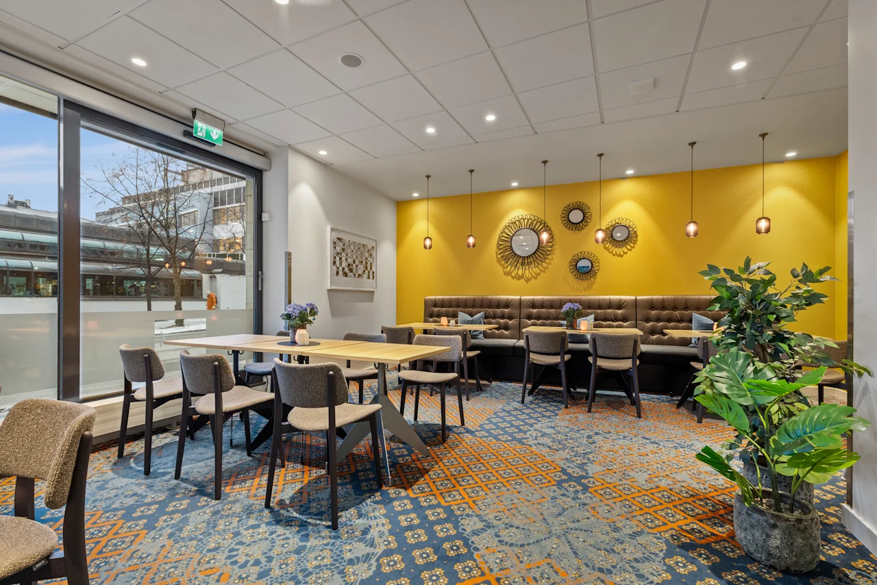 Ett färgglatt och inbjudande mat- eller loungeområde med gula väggar, mönstrad matta och gott om sittplatser. En utgångsskylt är synlig.