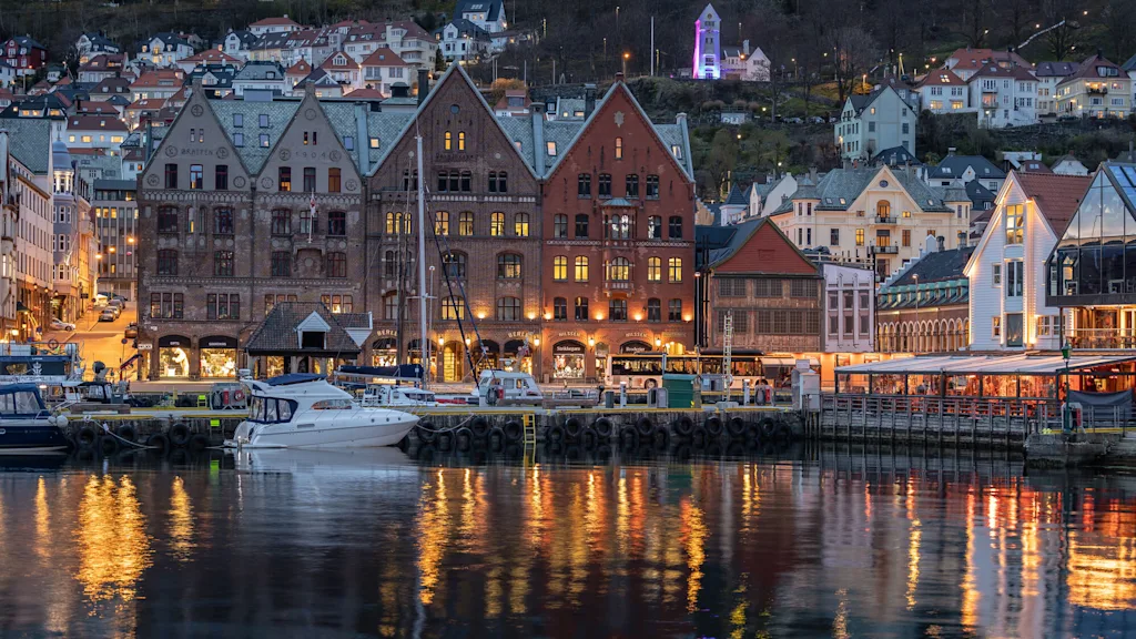 Historiska vattensidebyggnader speglas i vattnet, upplysta med varma ljus i skymningen. Båtar förtöjda i närheten, hus på en kulle och ett glödande torn i bakgrunden. Text inkluderar "Bratten", "1910", "Skagenkaien".
