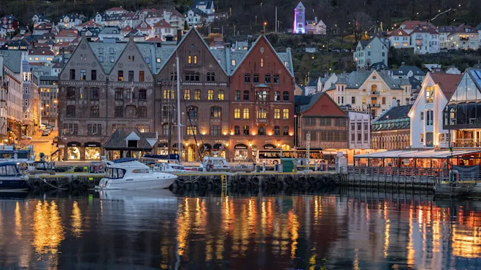 Historic waterfront buildings reflect on water, illuminated with warm lights at dusk. Boats docked nearby, hillside homes and a glowing tower in the background. Text includes “Bratten”, “1910”, “Skagenkaien”.