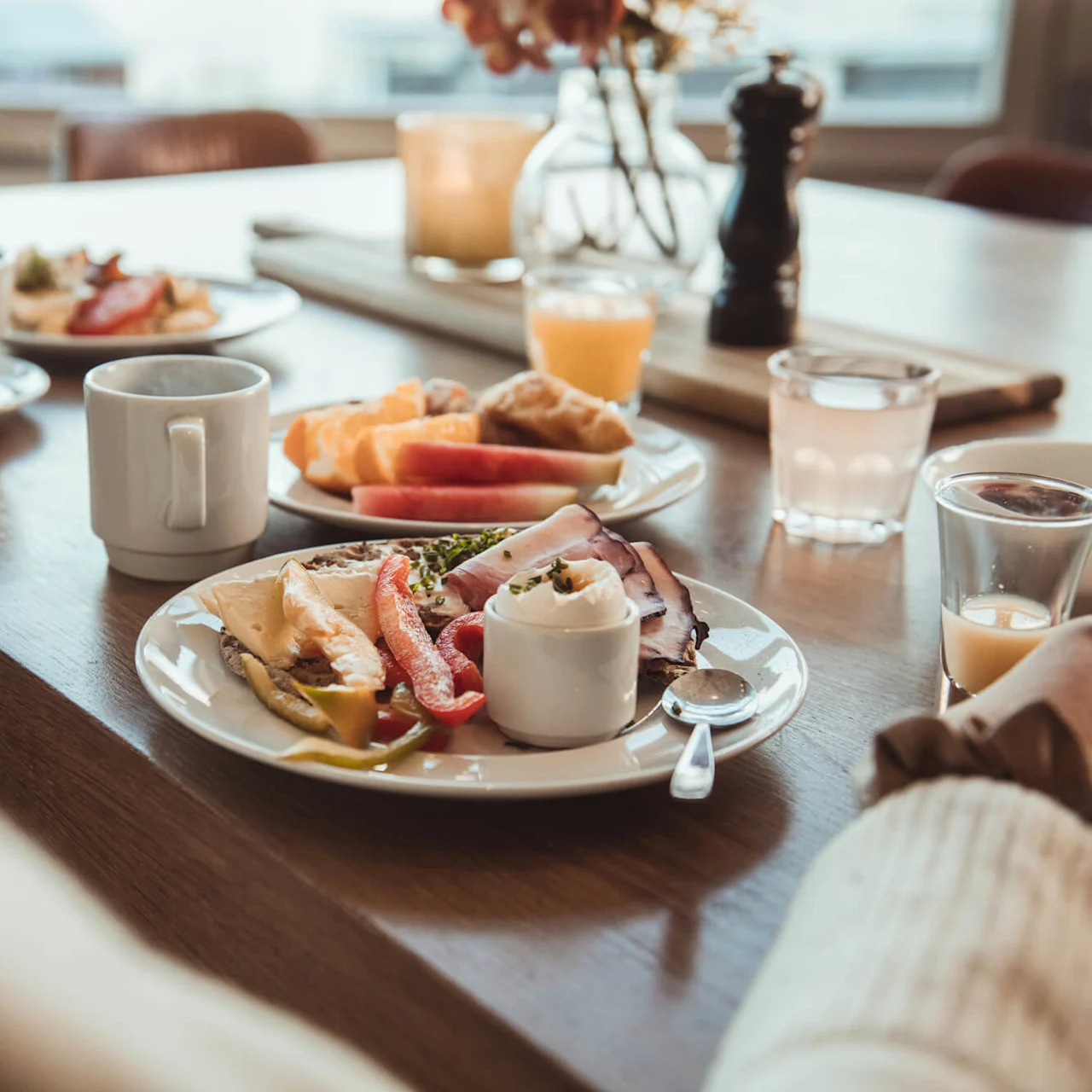 A breakfast spread with plates of ham, cheese, and vegetables. Cups of coffee and juice are on a wooden table in a softly lit dining room.