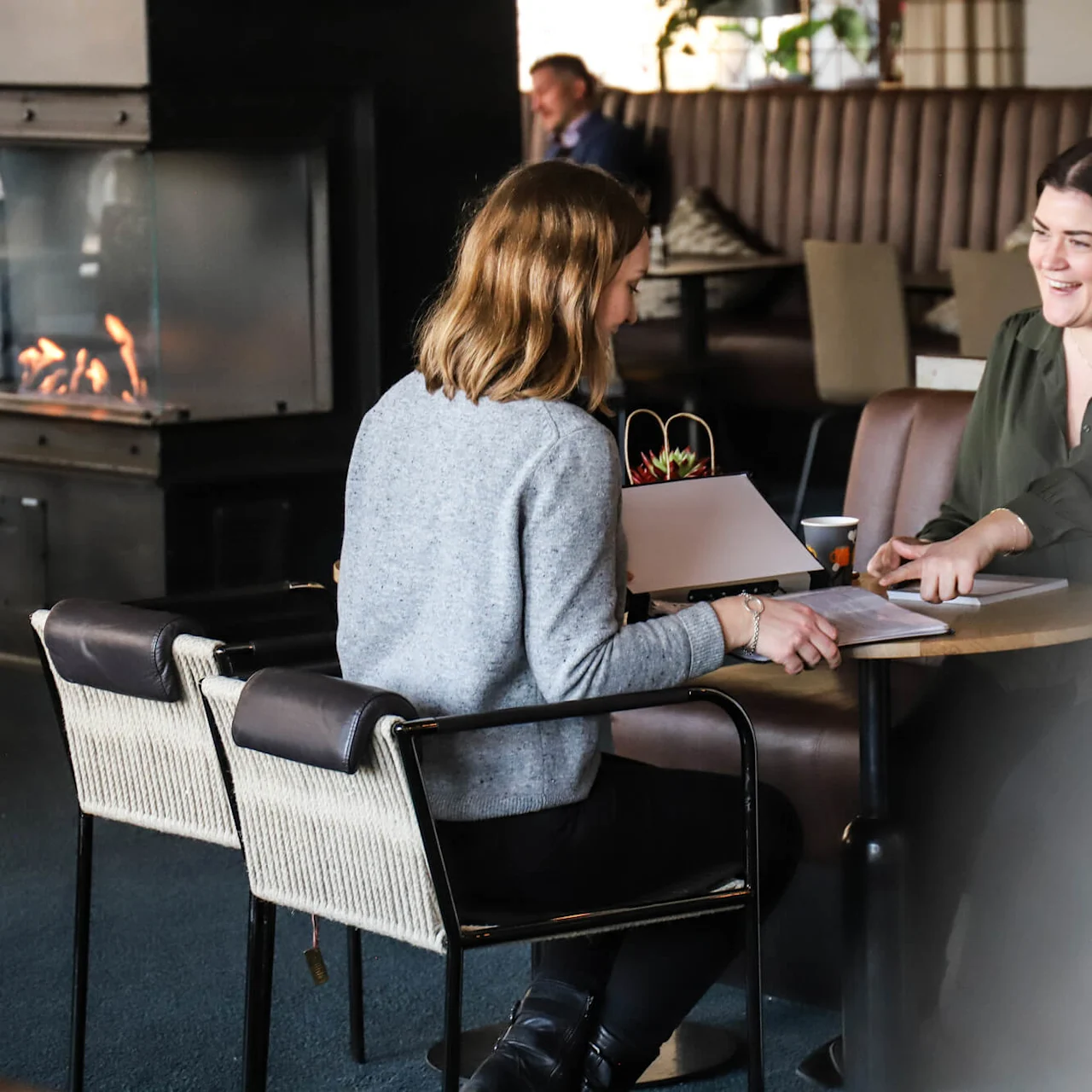 Två kvinnor sitter vid ett bord och diskuterar papper på ett mysigt café. En öppen spis värmer rummet, och en man sitter i bakgrunden.